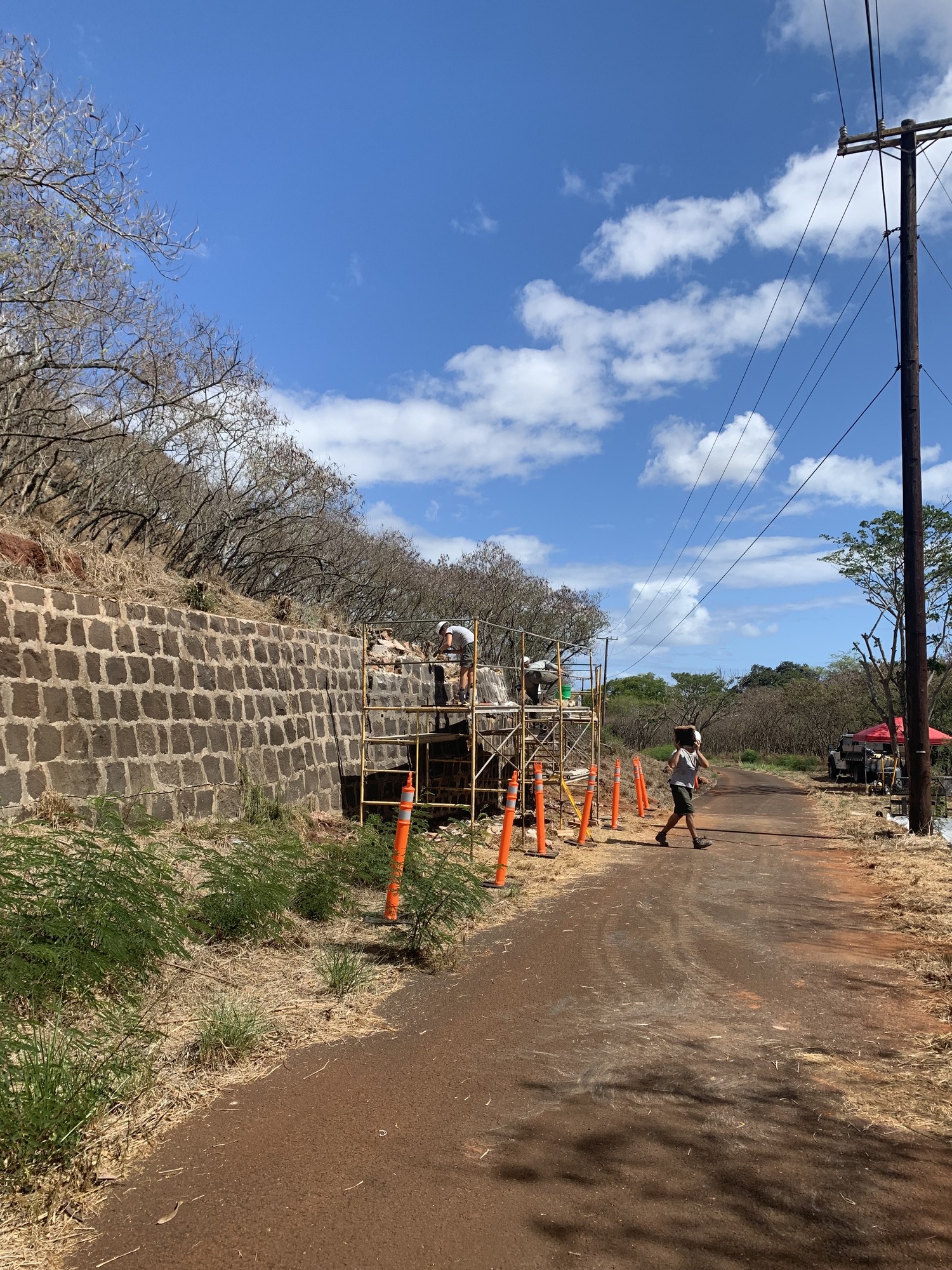 Two people on scaffolding repairing the rock wall and one person crossing the road adjacent to the wall.