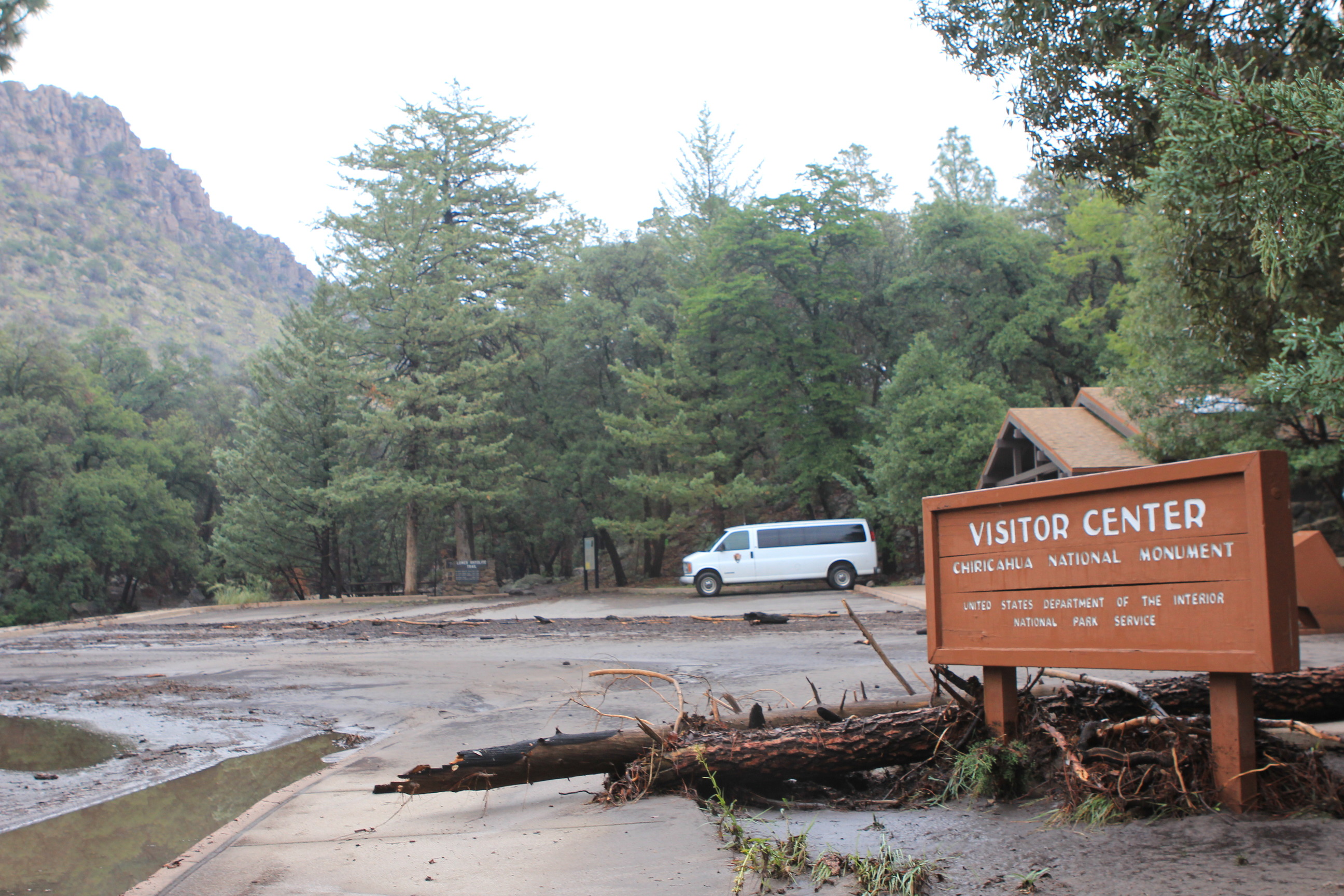 Silt, logs, and other debris pile against a visitor center sign and litter a parking lot with a white van parked in the far corner. 