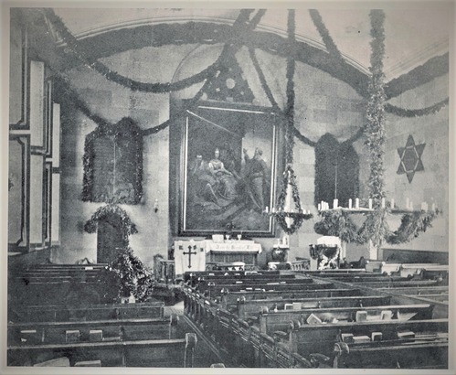 Image of a church interior, with pew benches, a large painting, wall tablets with inscriptions and various religious symbols. 