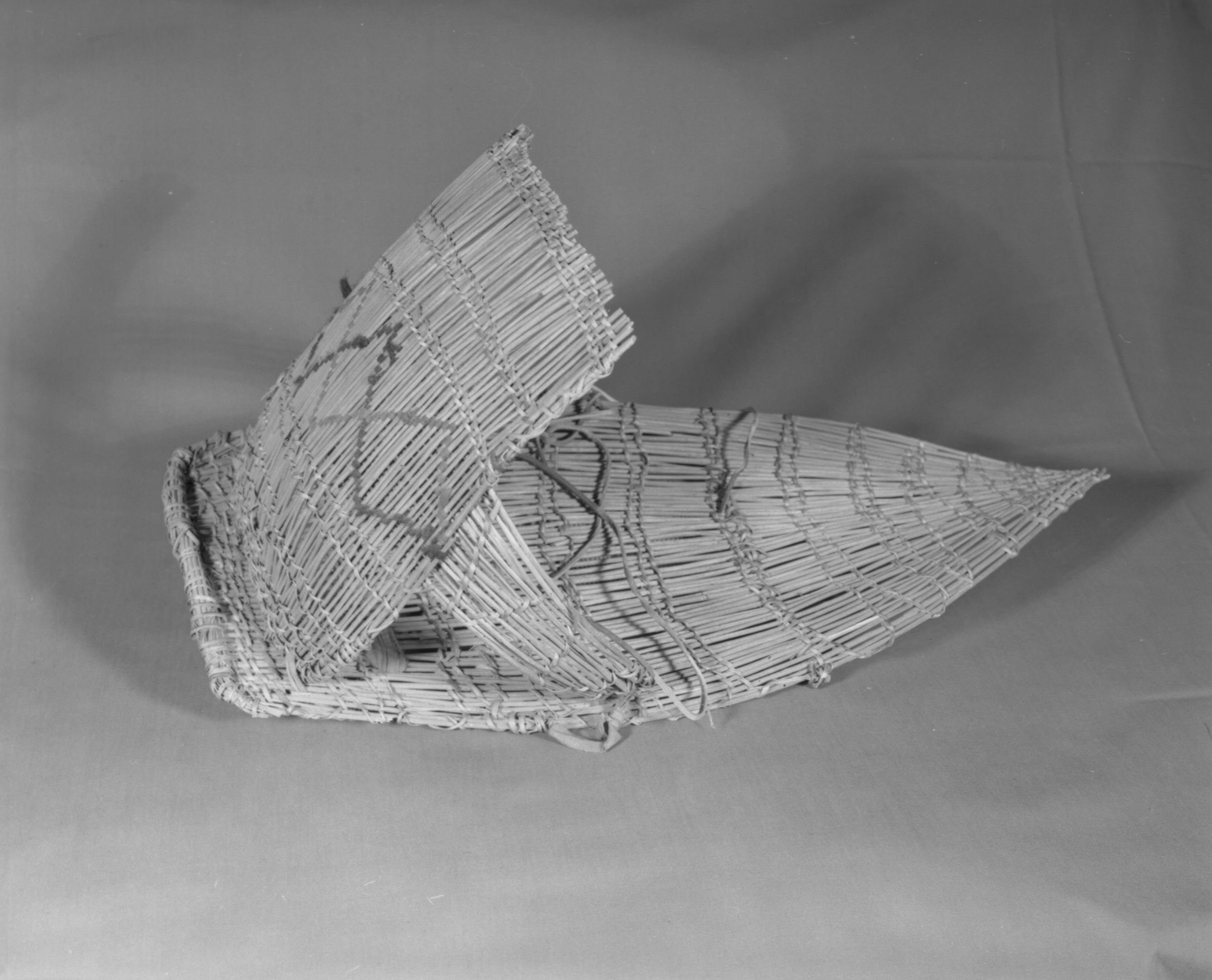 Private Collection. Object: Miwok cradle basket. Dimensions: 23 x 11 in center. Hood 12 x 9. Bows 7". Materials: Scraped willow shoots, splite ceanothus cuneatus, red & black wool yarn, mattress ticking, other unidentified cloth and commercial leather boot laces. Technology: Diagonally twined, up to the right slant of turns, splices are made by overlapping material. Hood has alternate rows slanting in opposite directions. Side bows have zigzagging rows of twining with loops on the ends that are bound. Collection History: From Mr. Halls collection in Sonora. Photographed by Michael Dixon. Catalogued by Craig Bates and Barbara Beroza. Yosemite Collections, National Park Service.