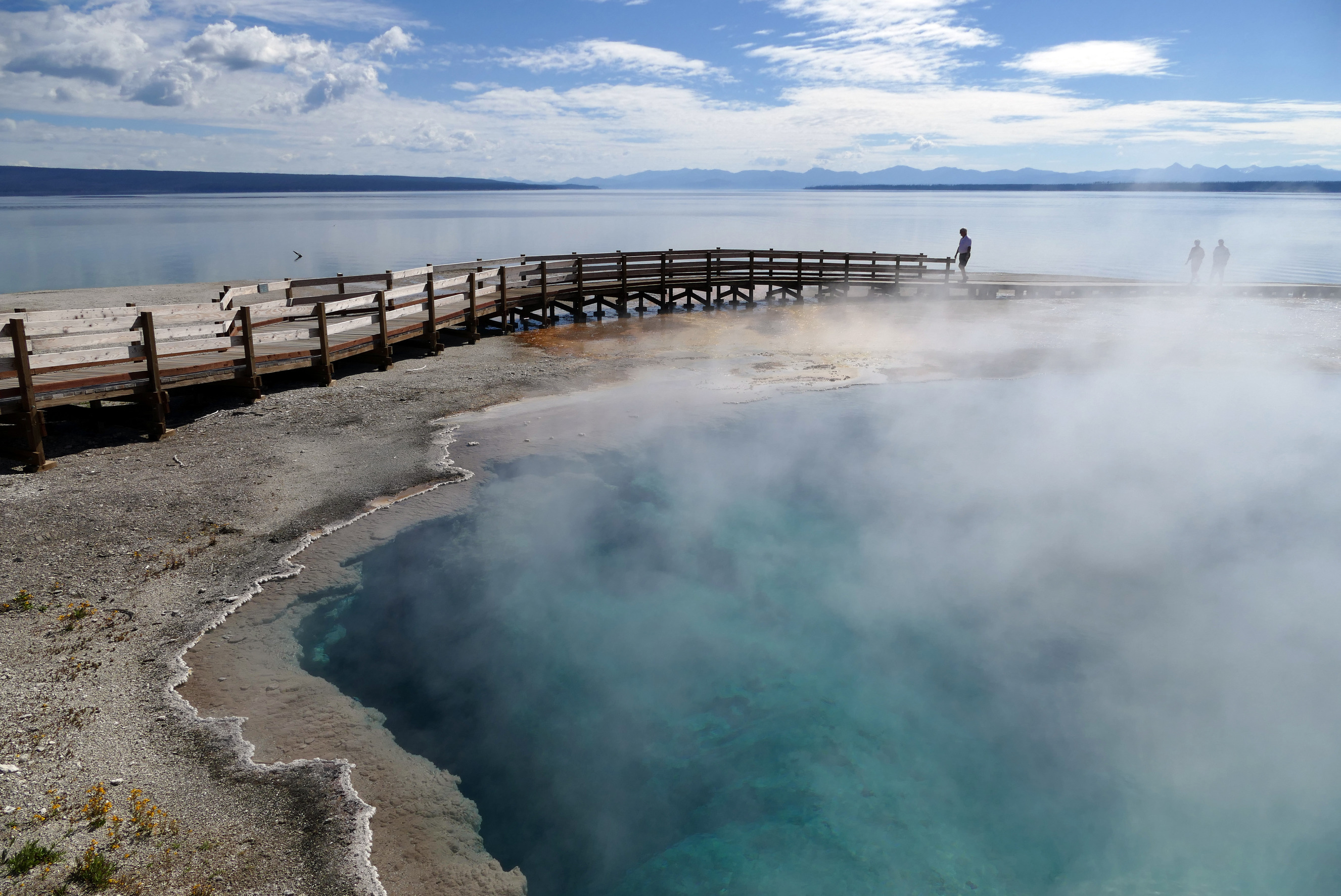 Looking out over a blue colored hot spring at a boardwalk and Yellowstone Lake behind that.