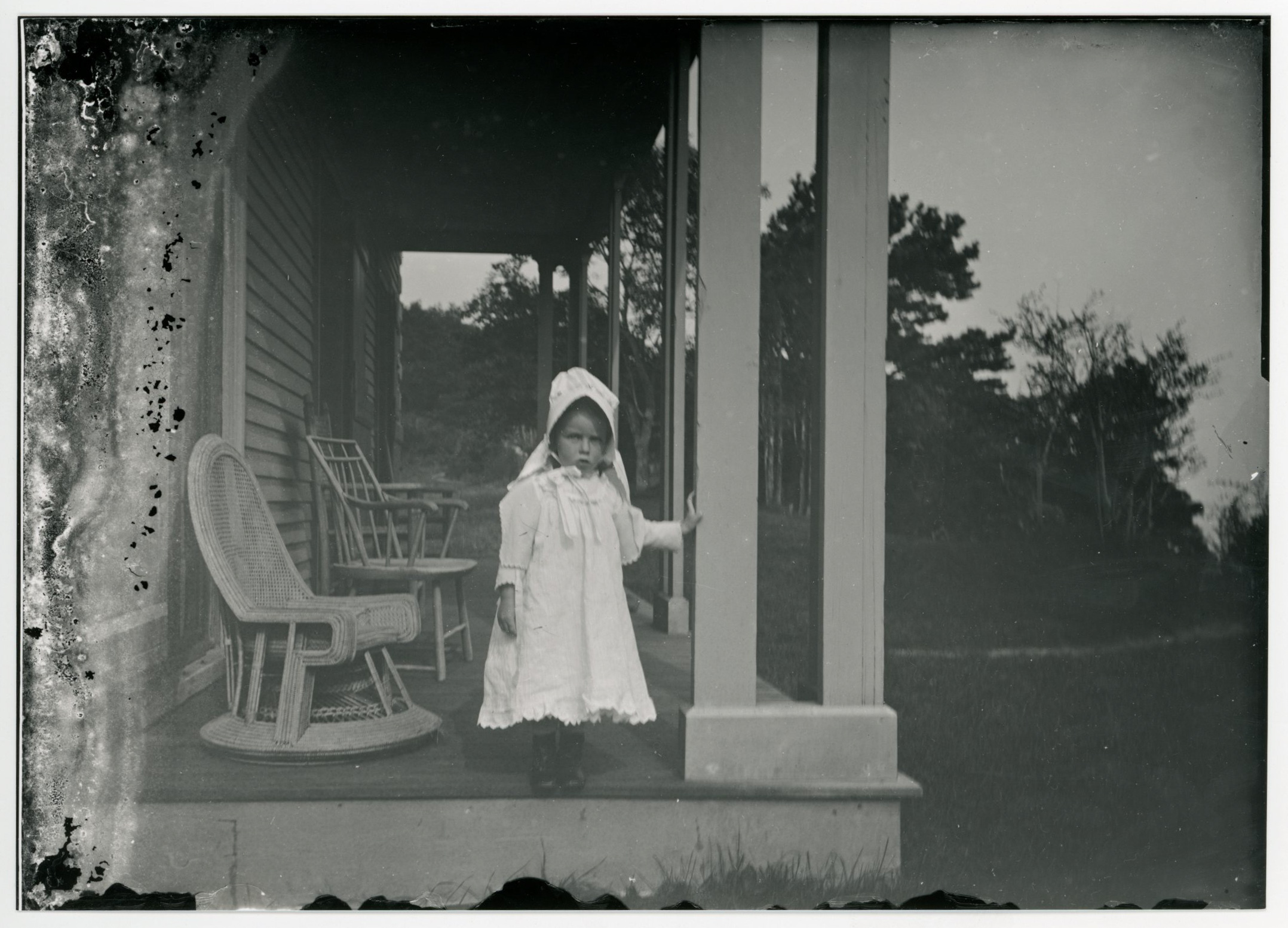 A young white girl wearing a large sunbonnet stands on a porch next to a wicker hair. Wood chair behind them. Trees in background.