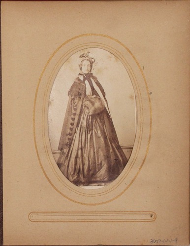 Black and white photograph of woman dressed in a shawl and bonnet and is carrying a fur muff.