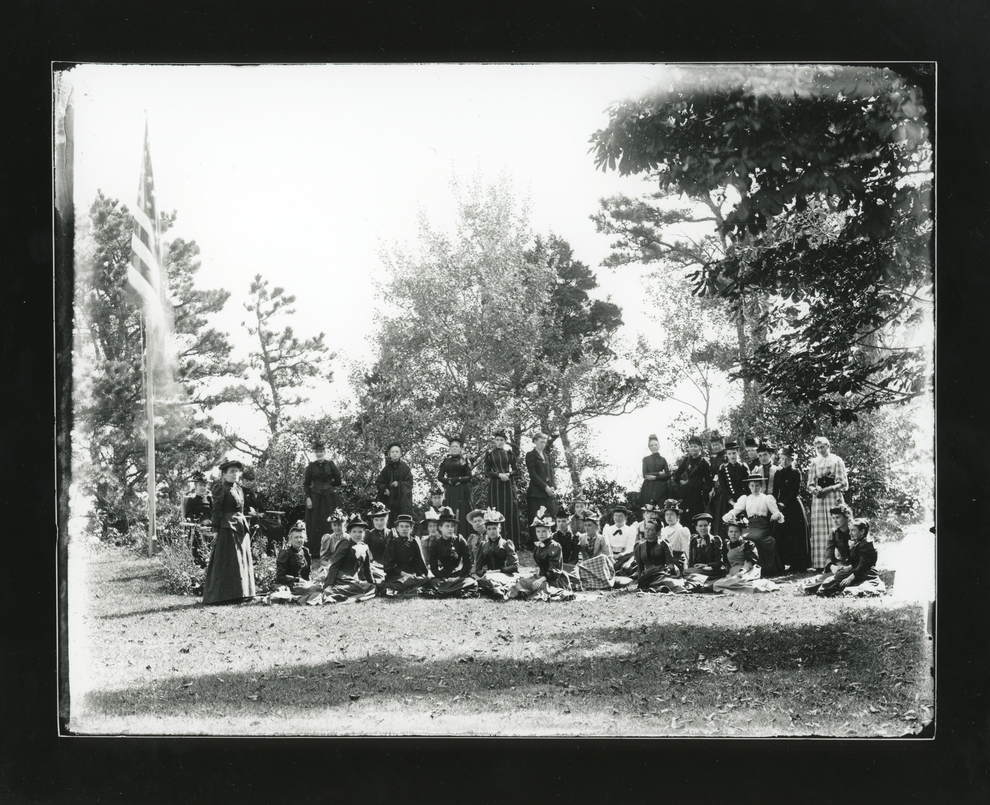 A large group of white women stand and sit outdoors in front of trees. All the women wear dresses and hats. An American flag flies at the left.