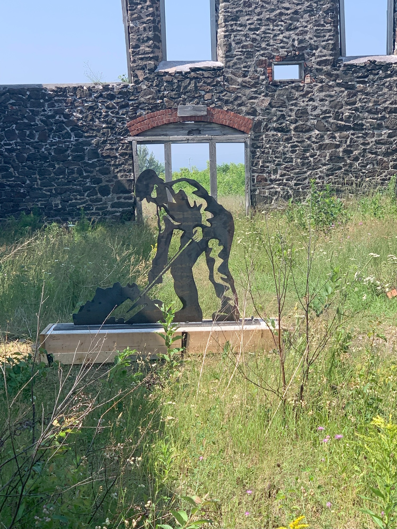 A metal sculpture of a man bent over a with his shovel in a pile of coal stands in the grass growing inside the ruins of an old mine building. 