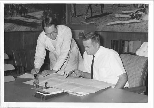 One individual sits at a desk while another looks over his shoulder at a document sitting on the desk in front of them. 
