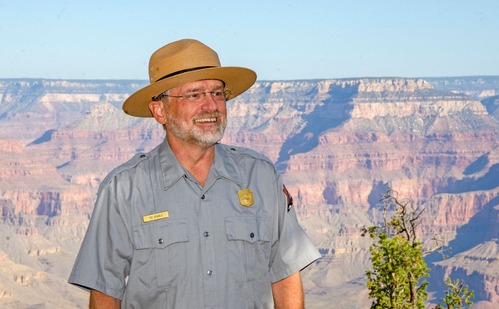 A National Park Service ranger in uniform and a wide-brimmed hat stands smiling in front of the Grand Canyon landscape under a clear sky.