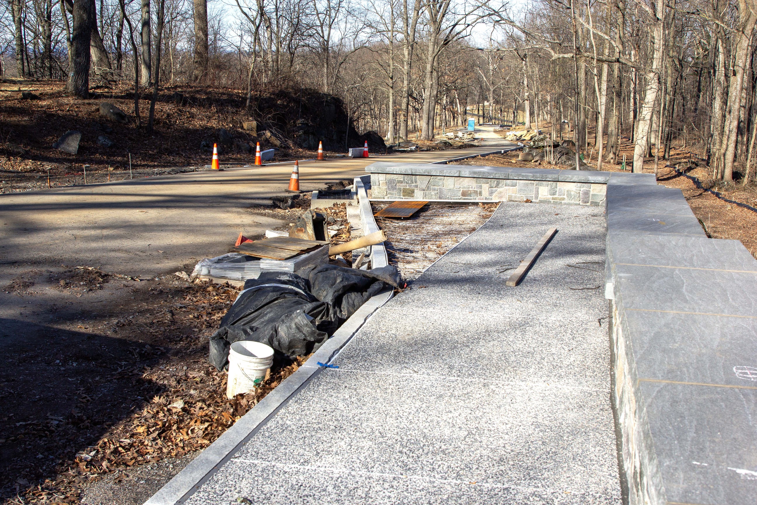 A sidewalk along a stone wall appears in the right of the photo. Construction materials and orange construction cones are in the left half of the photo. 