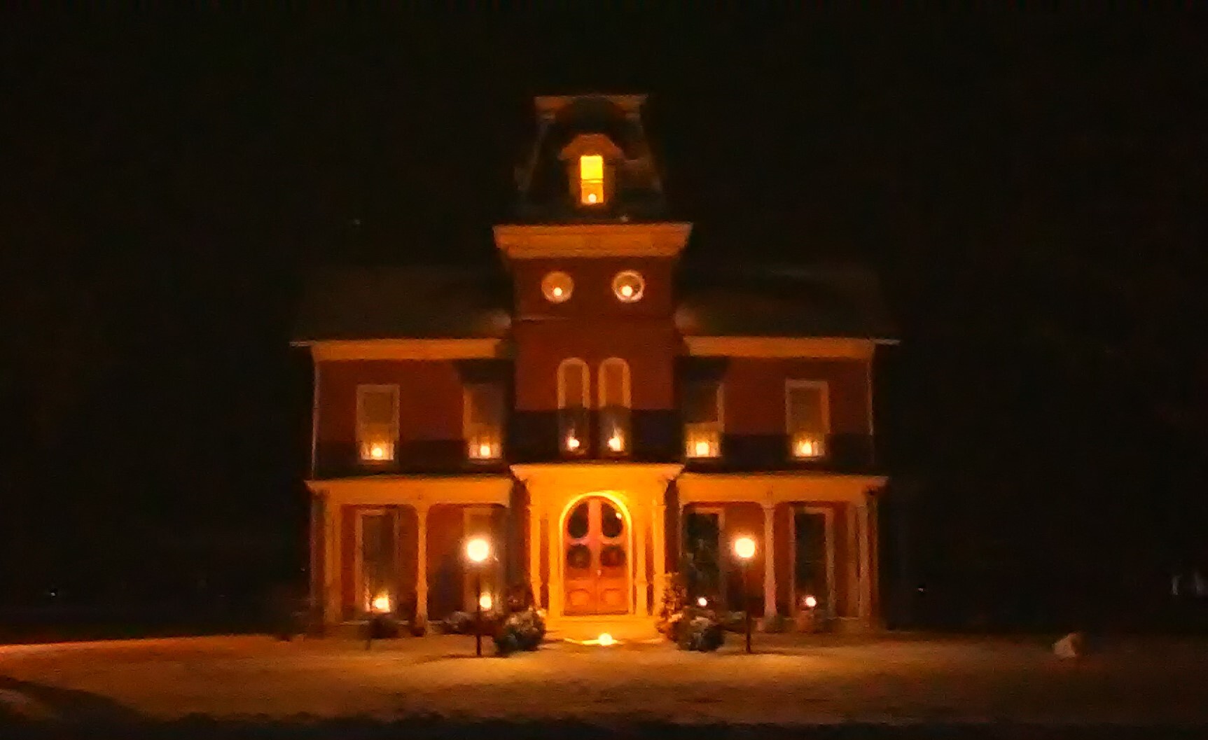 This color photograph was taken at night time. It depicts a second empire home, with two stories, and multiple windows. Each window has a candle lit inside. The candle light and two gas lamps outside illuminate the structure.