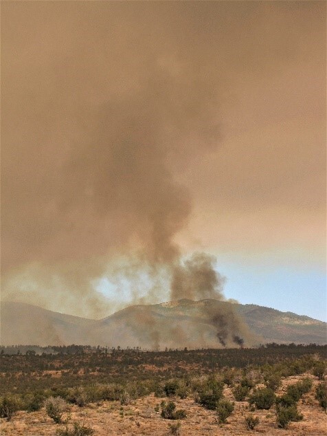 A cloud of brown-gray smoke covers the sky over the mountains and the shrub-covered mesas