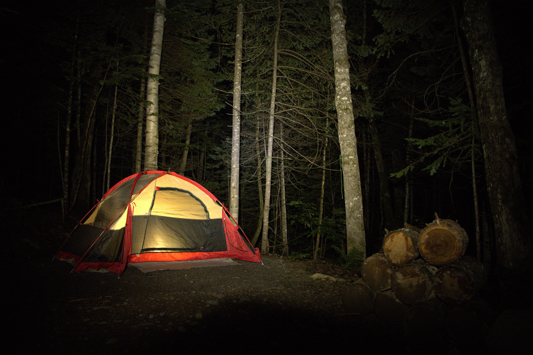 At night, a red tent is illuminated with warm light from the inside. The tent is surrounded by tall trees and a stack of firewood rests next to the tent. 