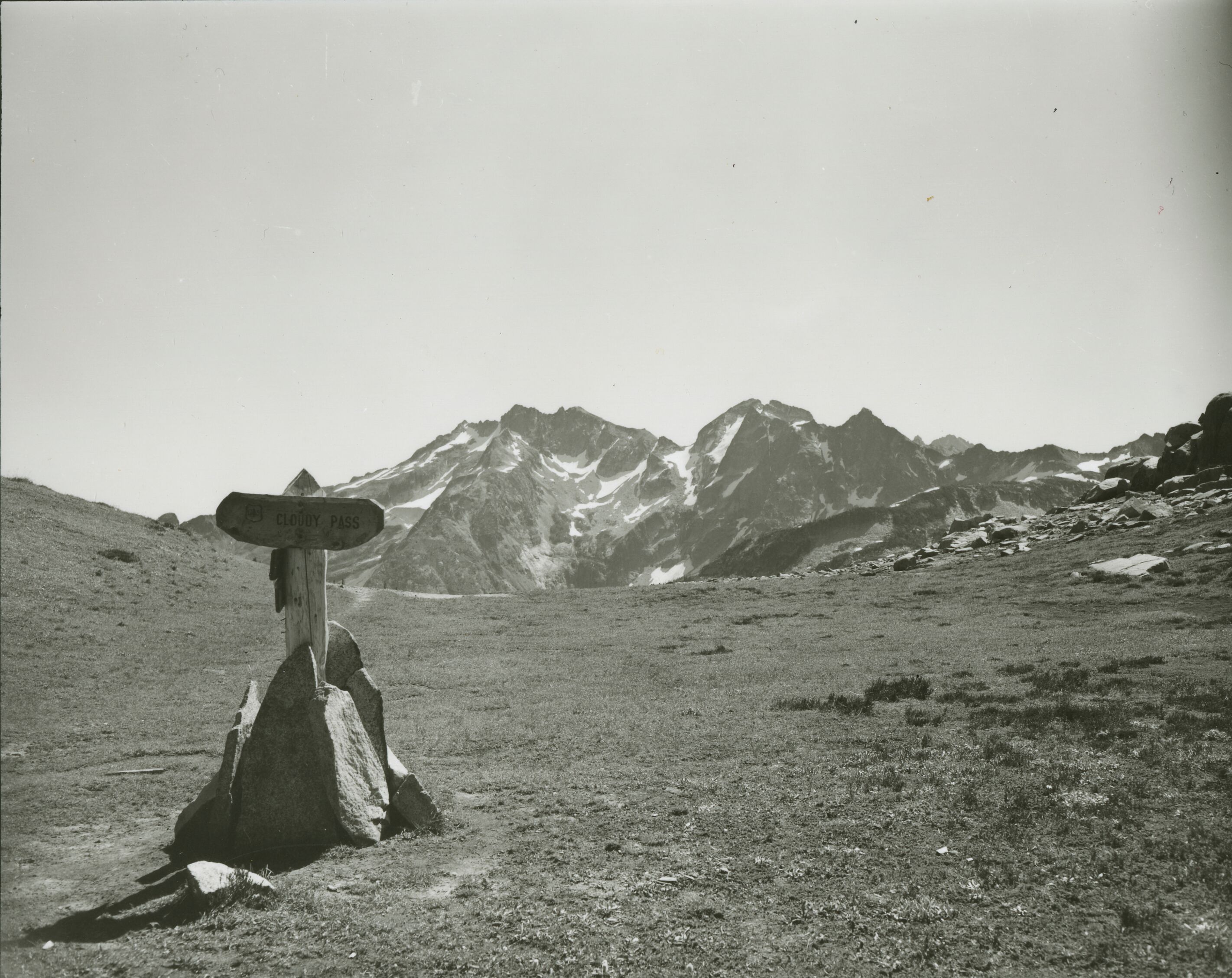 A wooden sign that says 'Cloudy Pass' on a high mountain clearing. Mountain peaks in the background.