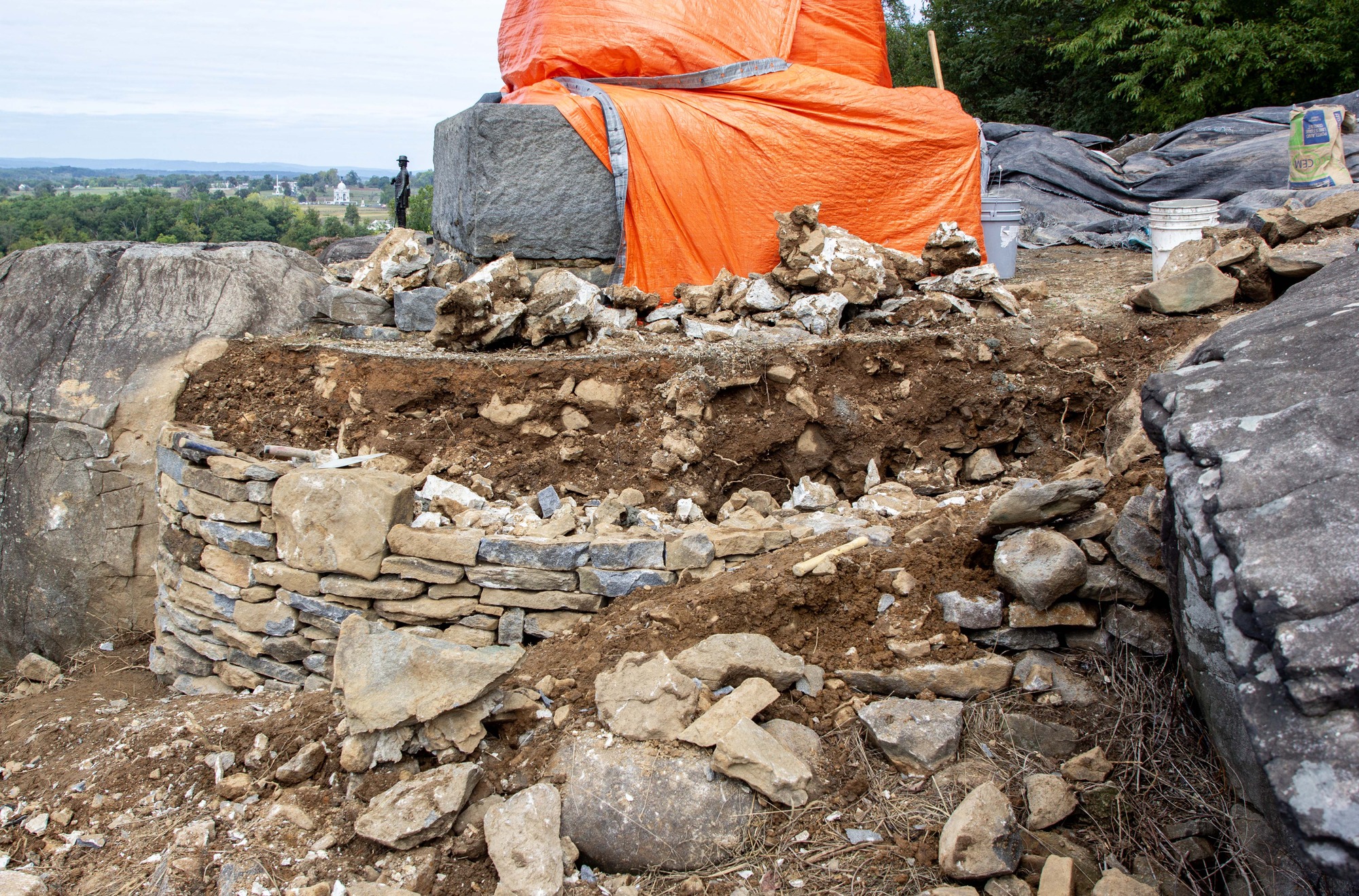 A stone wall is being built to the center of a two very large boulders. In the center is a tall grey stone monument with a bright orange tarp wrapped around the bottom of the structure.