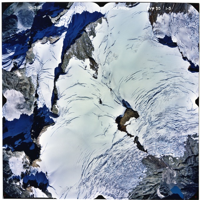 Color aerial photograph of glacier and alpine lake on mountain peak.