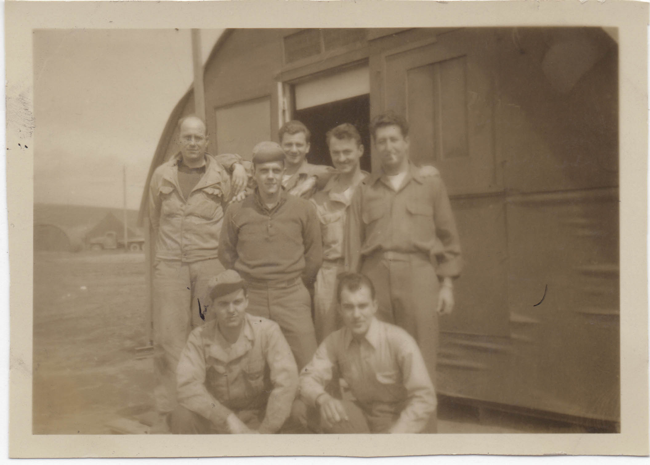 Seven men pose in front of a quonset hut
