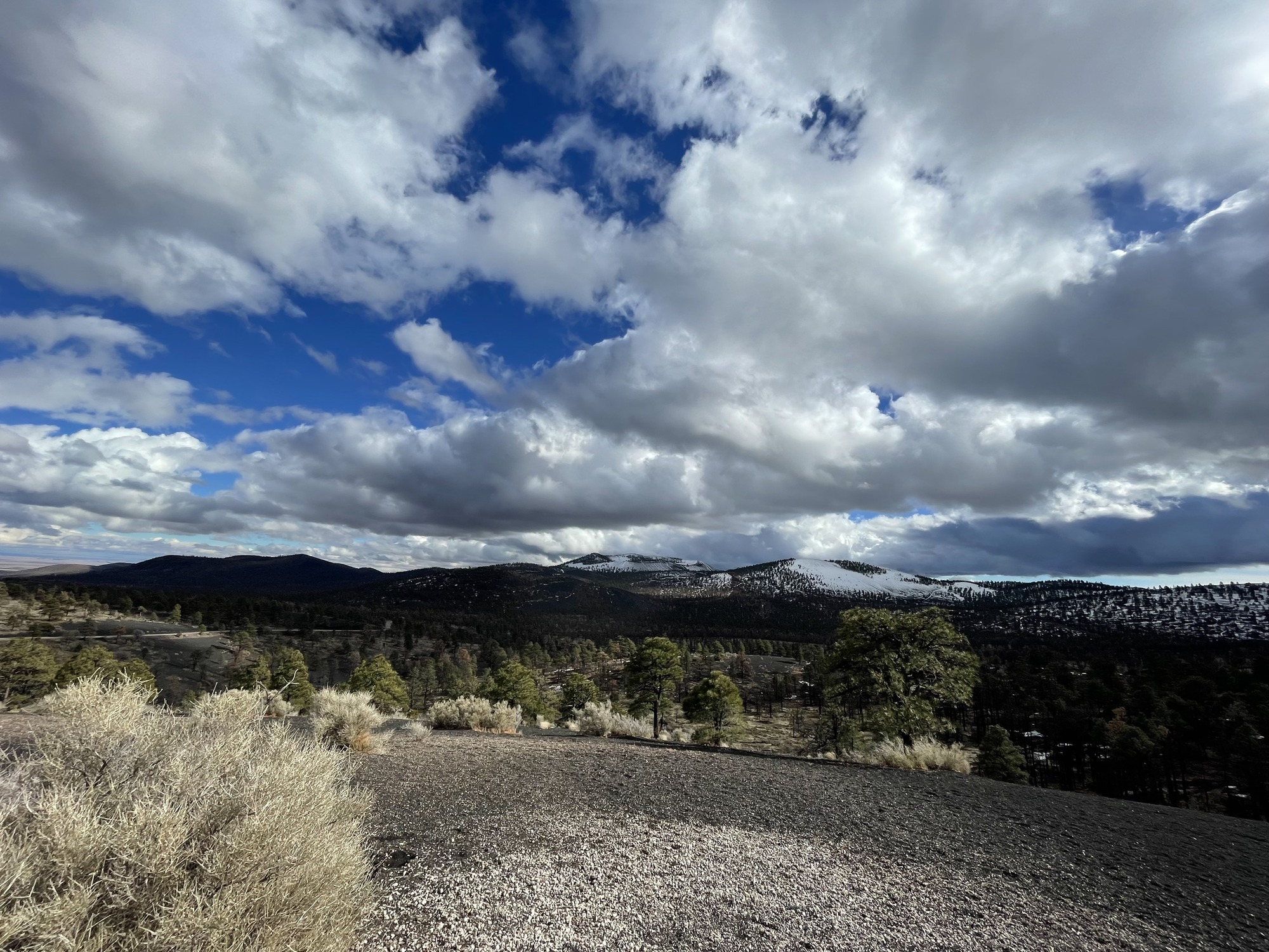 A wide view of rolling volcanic hills partially covered in snow surrounded by sparse pine trees and a blue sky with puffy white clouds. 