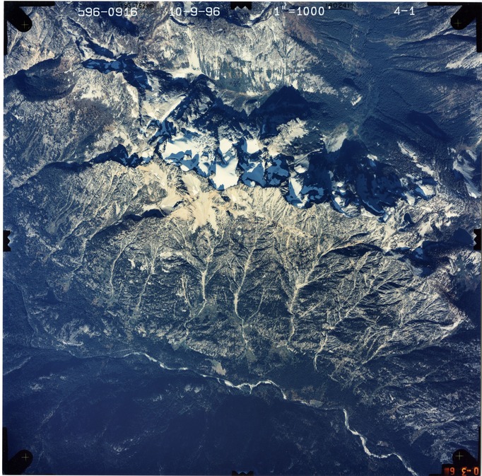 Color aerial photograph of glacier and snowfields on mountain peak with forested valleys below.
