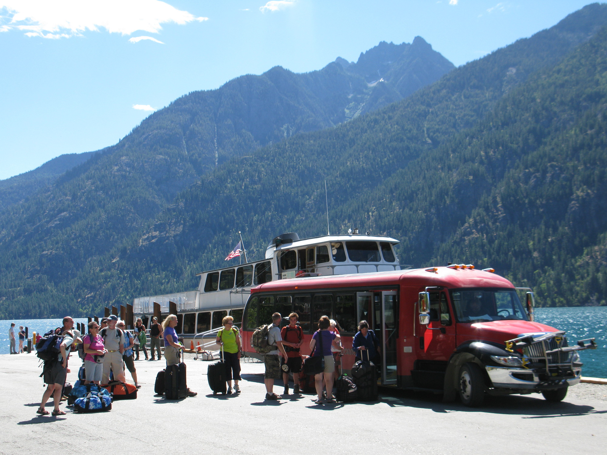 A group of fifteen people with bags wait to board a bus by a large lake and ferry boat.