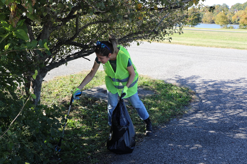 A person wearing a neon yellow safety vest, blue jeans, and gloves uses a litter picker to collect trash into a black garbage bag next under a tree in a parking lot. Trees and green foliage are visible, and in the background, a body of water and a grassy field can be seen under a clear sky.