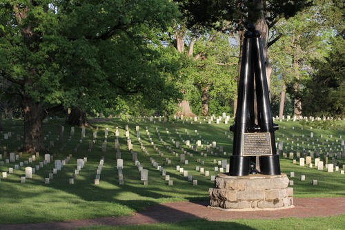 Three black inverted cannon tubes mounted on a stone base with headstones in the background. 
