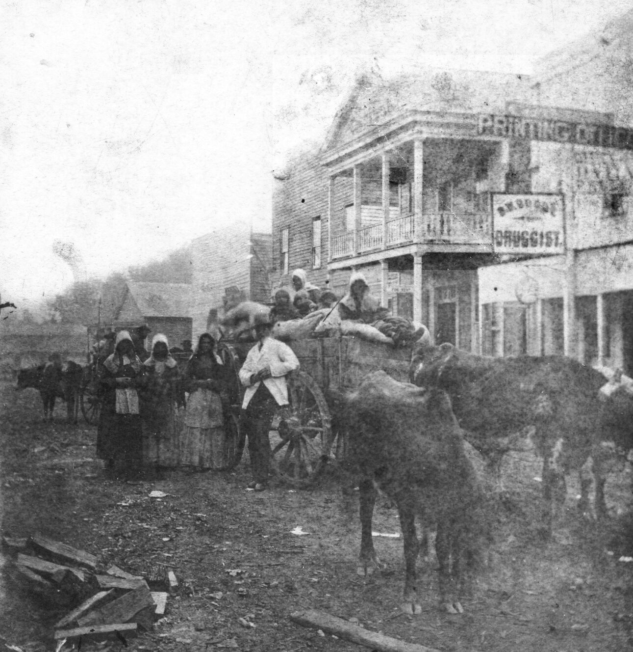 Women in bonnets and a man in a white coat stand by a wagon filled with what appears to be bags of grain and pulled by two ox, in front of the old Hot Springs Hotel.