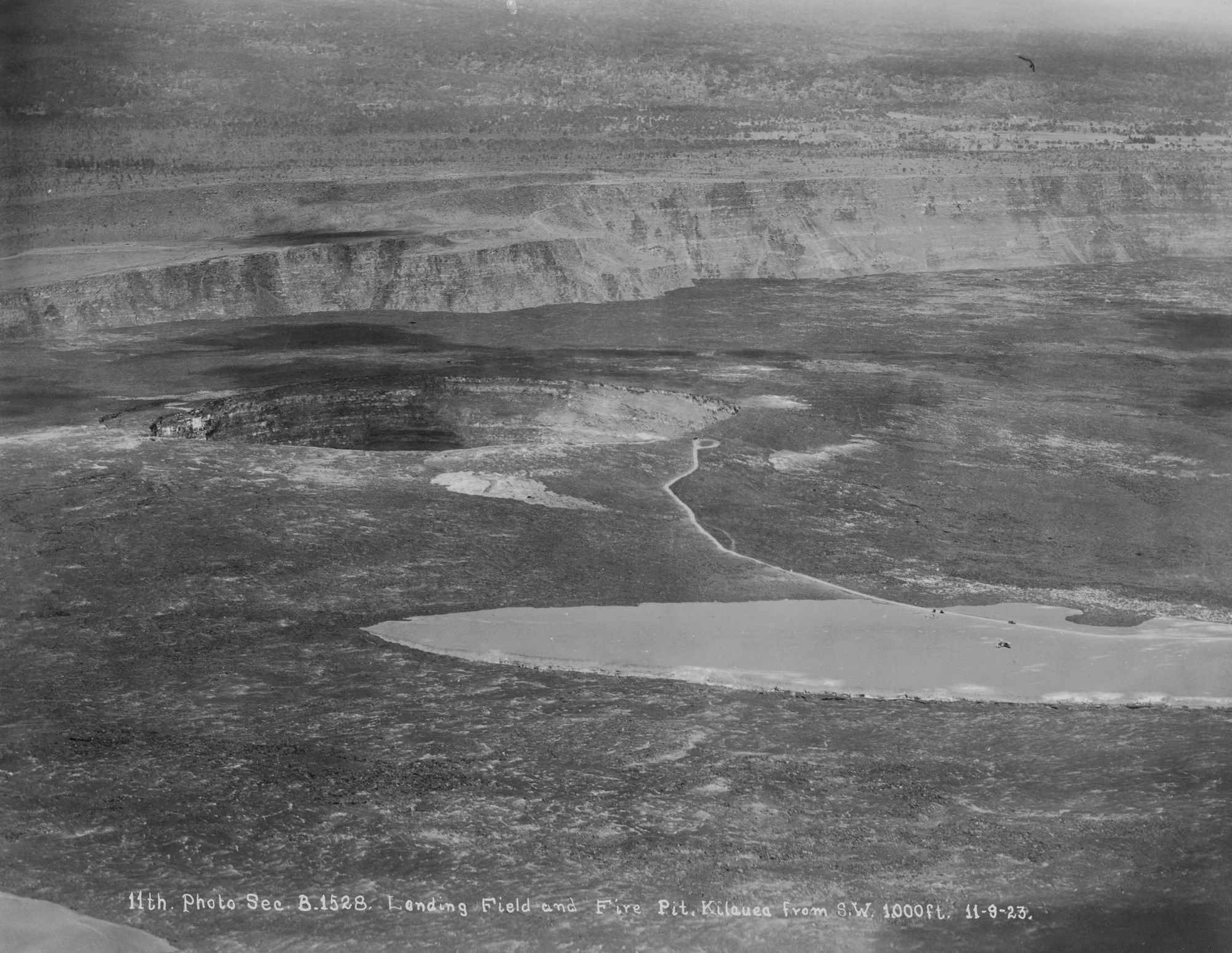 Black and white photograph of a landscape taken at a high vantage point. To the right of the image, an oval-shaped piece of flat, smooth ground interjects the rough, rocky landscape. A thin white road is seen travelling through the smooth ground and extending further towards the background, ending in a cul-de-sac. The end of the road sits near the rim of a Halemaʻumaʻu crater. The calderaʻs wall begins just beyond the crater. Handwriting at the bottom reads, “11th Photo Sec. B1528. Landing Field and Fire Pit, Kilauea from S.W. 1000 ft. 11-9-23.”