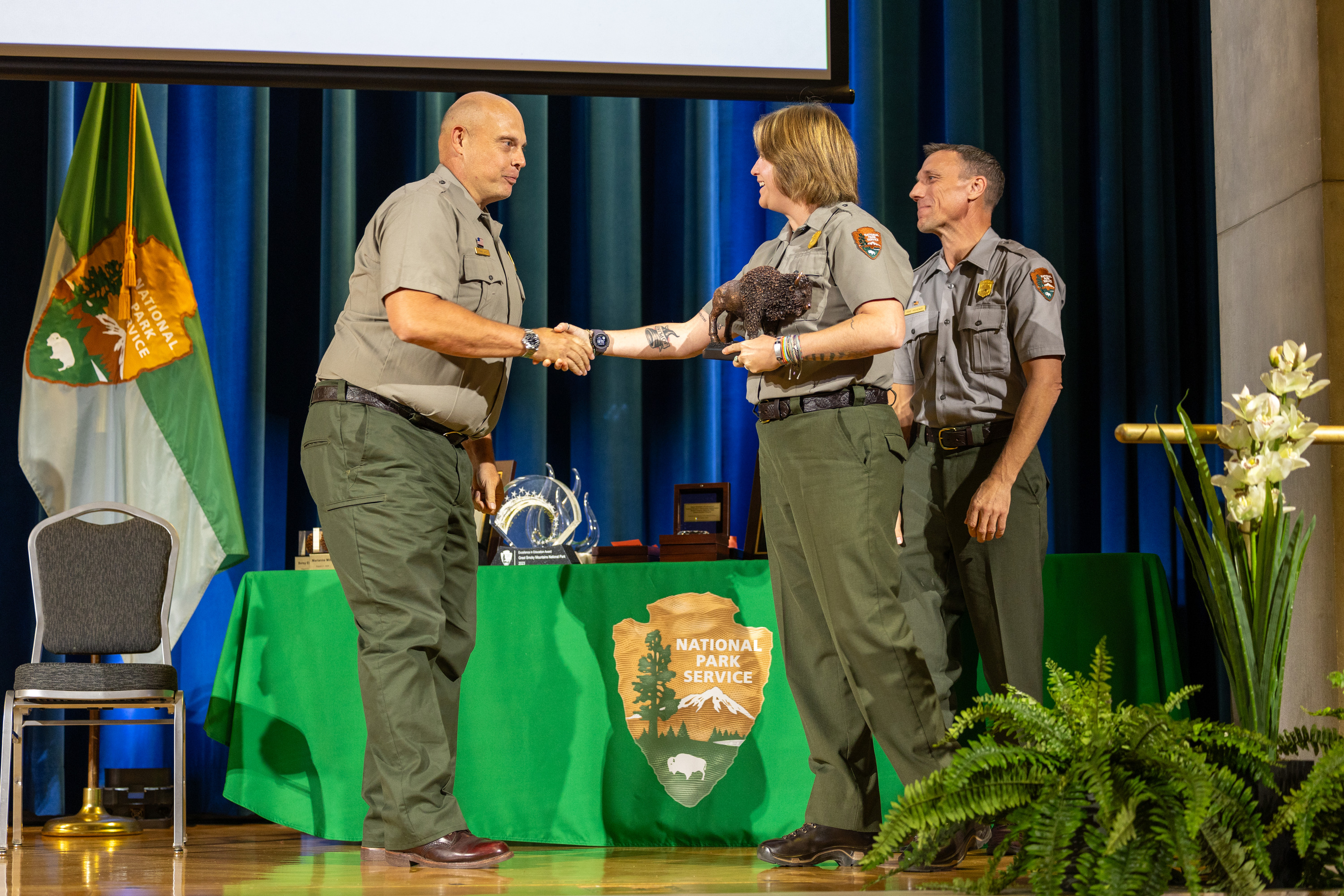 A female award recipient and two men in green and gray park ranger uniforms at an awards ceremony.