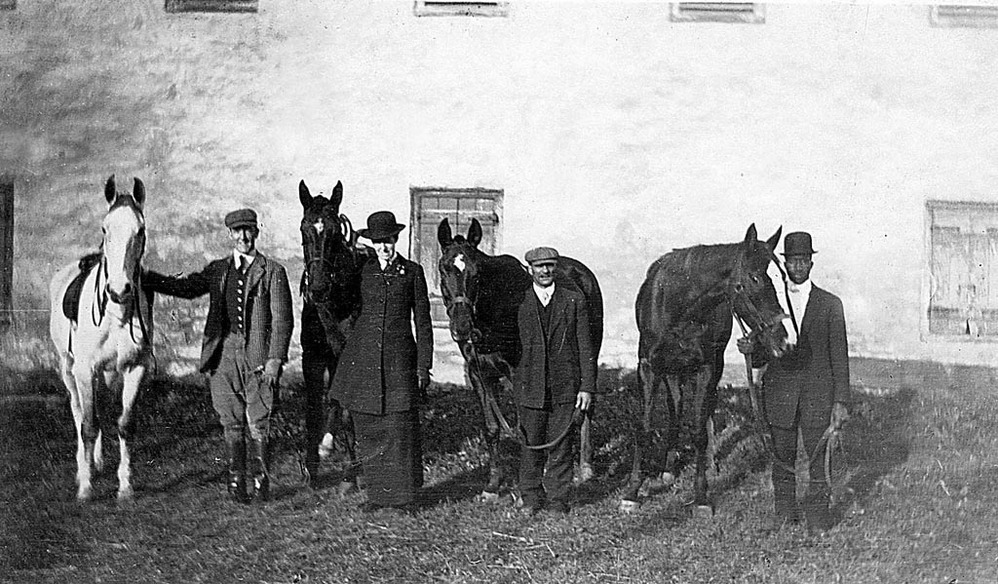 A group of four people stand with four horses in front of a stone building.