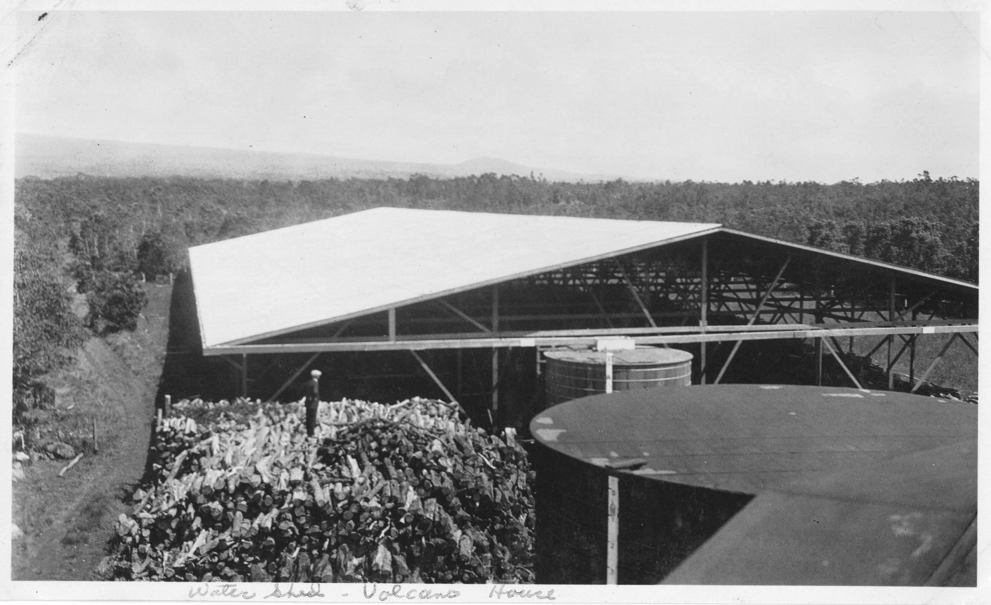 The image is in black and white. In the foreground of the image, there is a slightly blurry structure in the lower right corner. Behind this structure is a large, cylindrical, metal tank. To the left and slightly behind the tank is a giant pile of cut tree trunks. The pile is as wide as the tank. A man stands on top of the pile, looking to a shed behind the tank and pile. Another smaller, lighter-colored tank sits directly in front of the shed's center. The shed itself is quite large, spanning nearly the entirety of the photo from one end to the other. The roof is bright. None of the structure is enclosed. Posts and beams support the roof and ground the building. A dirt road runs alongside the wood pile and shed, running into the forest behind the shed. Dense forest surrounds the shed and continues into the background. The slope of a mountain is faintly visible in the distance. A label written on the bottom white border of the photo reads, "Water shed, Volcano House."