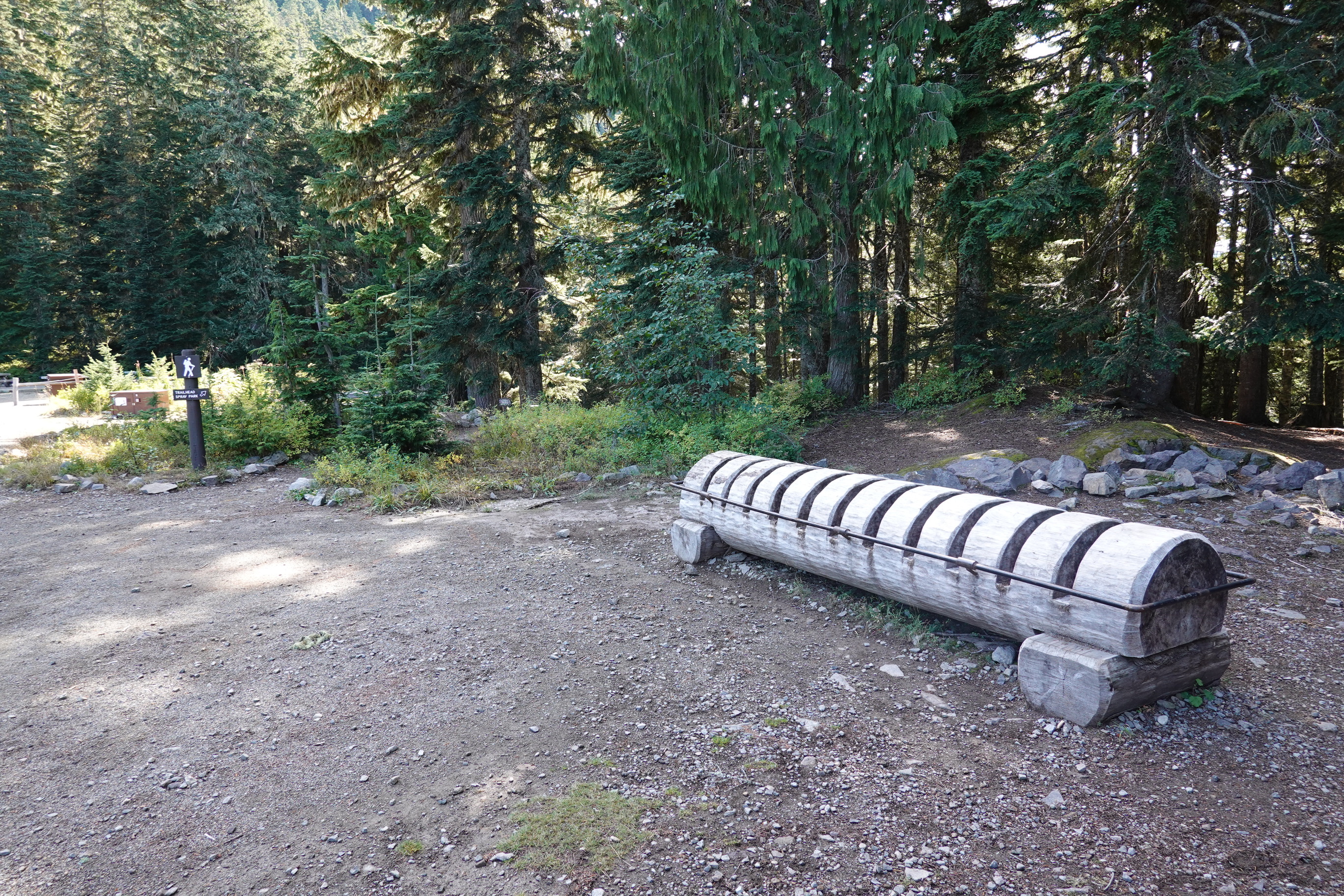 A large log with deep notches carved halfway into it and wrapped in a metal bar serves as a bike rack next to a trailhead. 