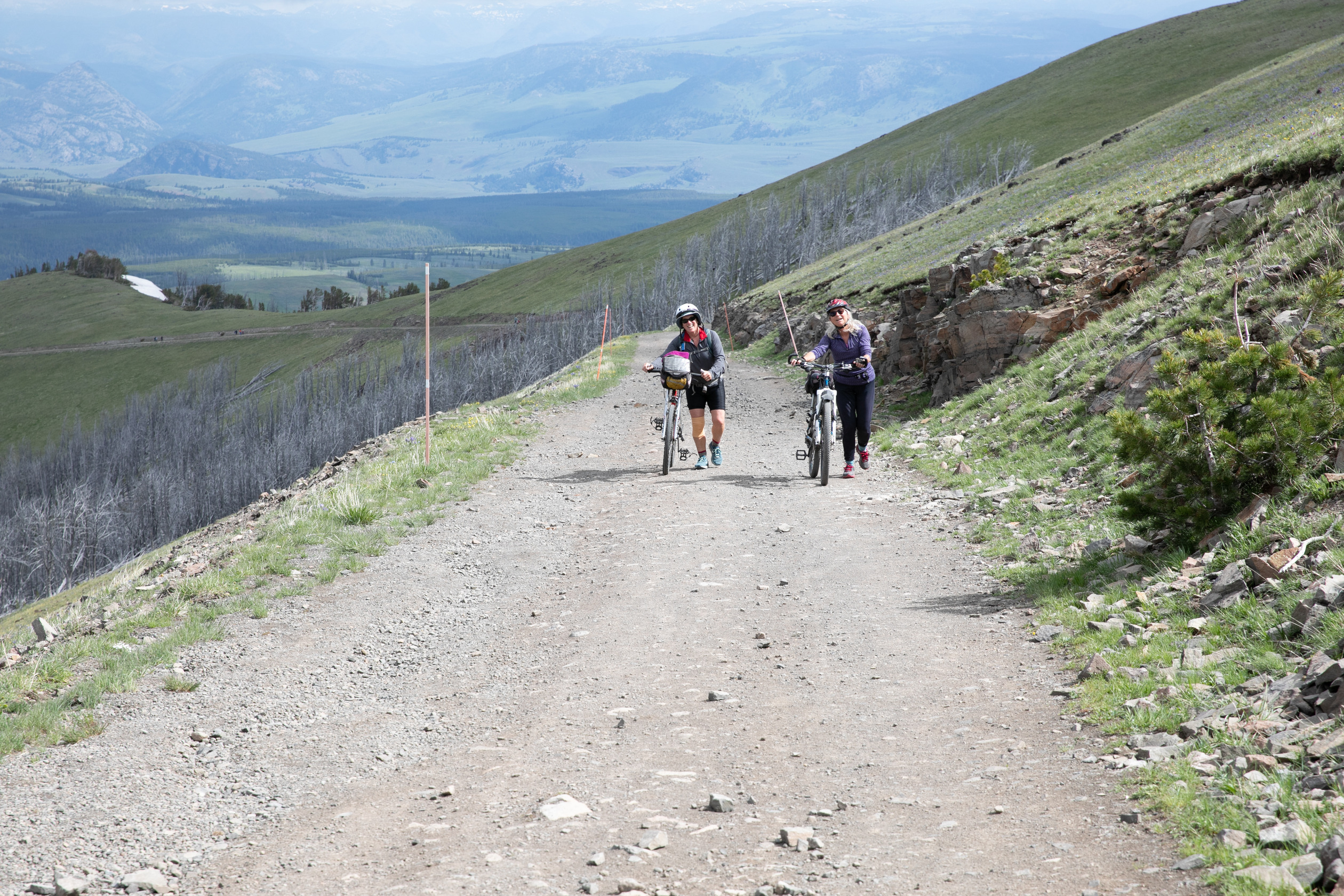 Two women walk their bikes up a dirt road on a mountain