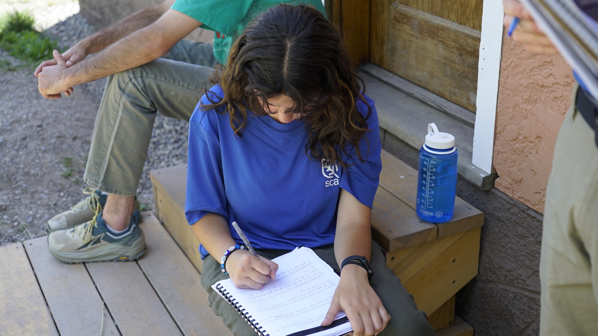 A woman with brown hair sits on a step to write in a notebook 