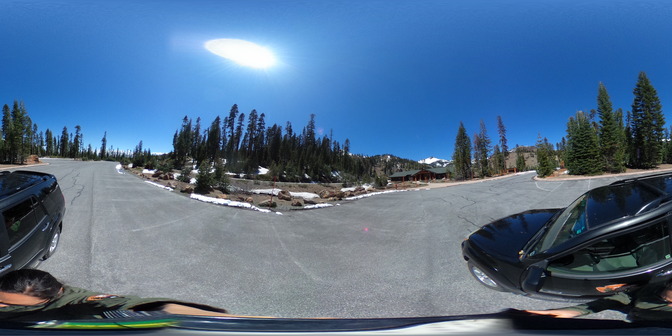 A 360-degree photo of a parking area lined by green conifer trees. A one-story building sits at one end below a snow-covered mountain.