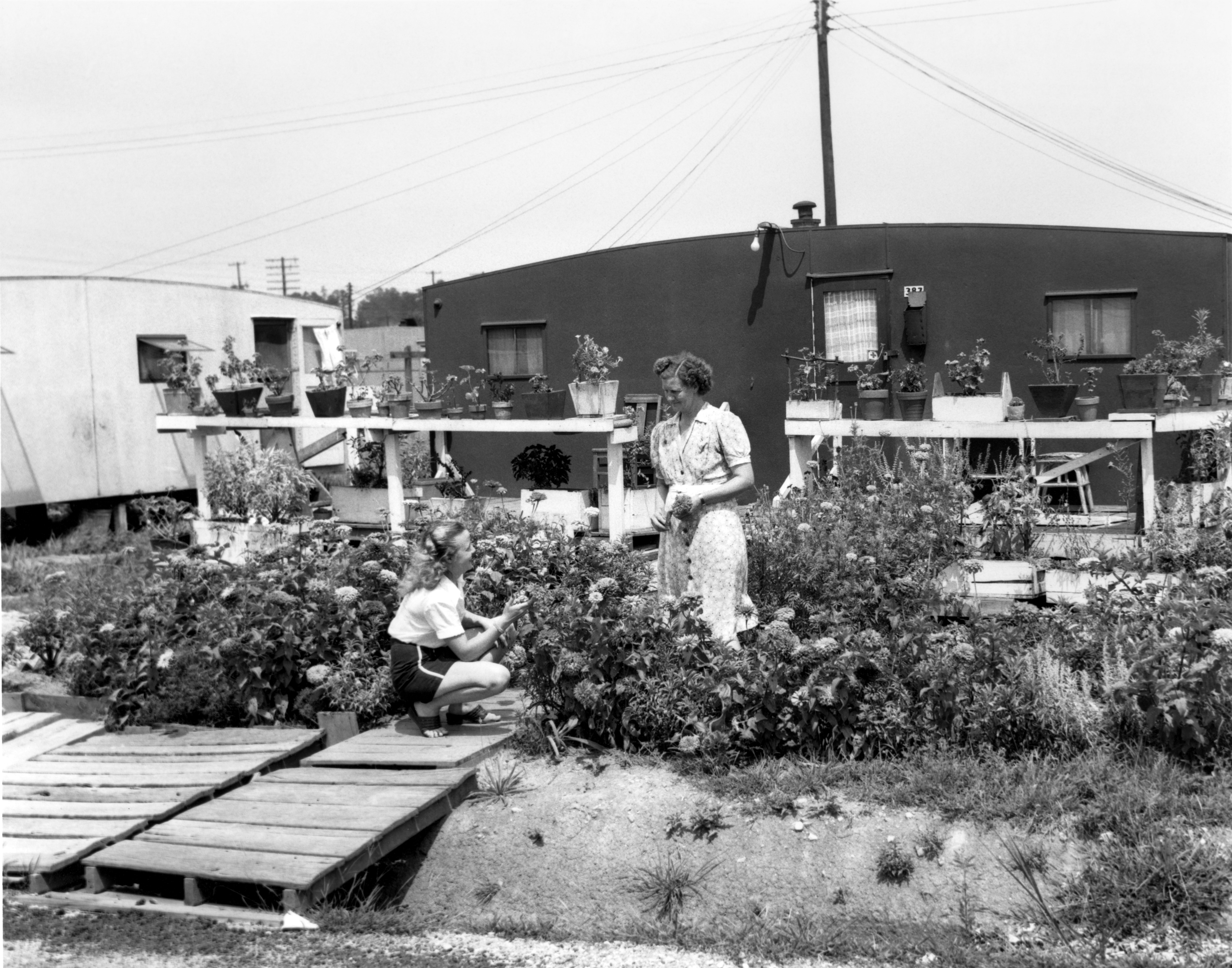 Black and white photo of two women tending to a garden in front of a trailer. 