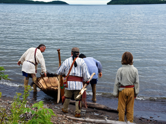 Four people in historic clothing grouped around the bison hide boat. Two are carrying it to the water.