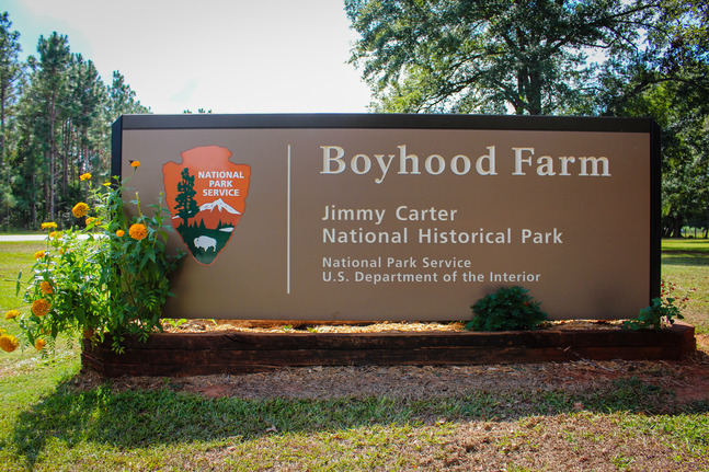 National Park Service sign surrounded by marigold flowers. 
Text: Boyhood Farm, Jimmy Carter National Historical Park, National Park Service, U.S. Department of the Interior 