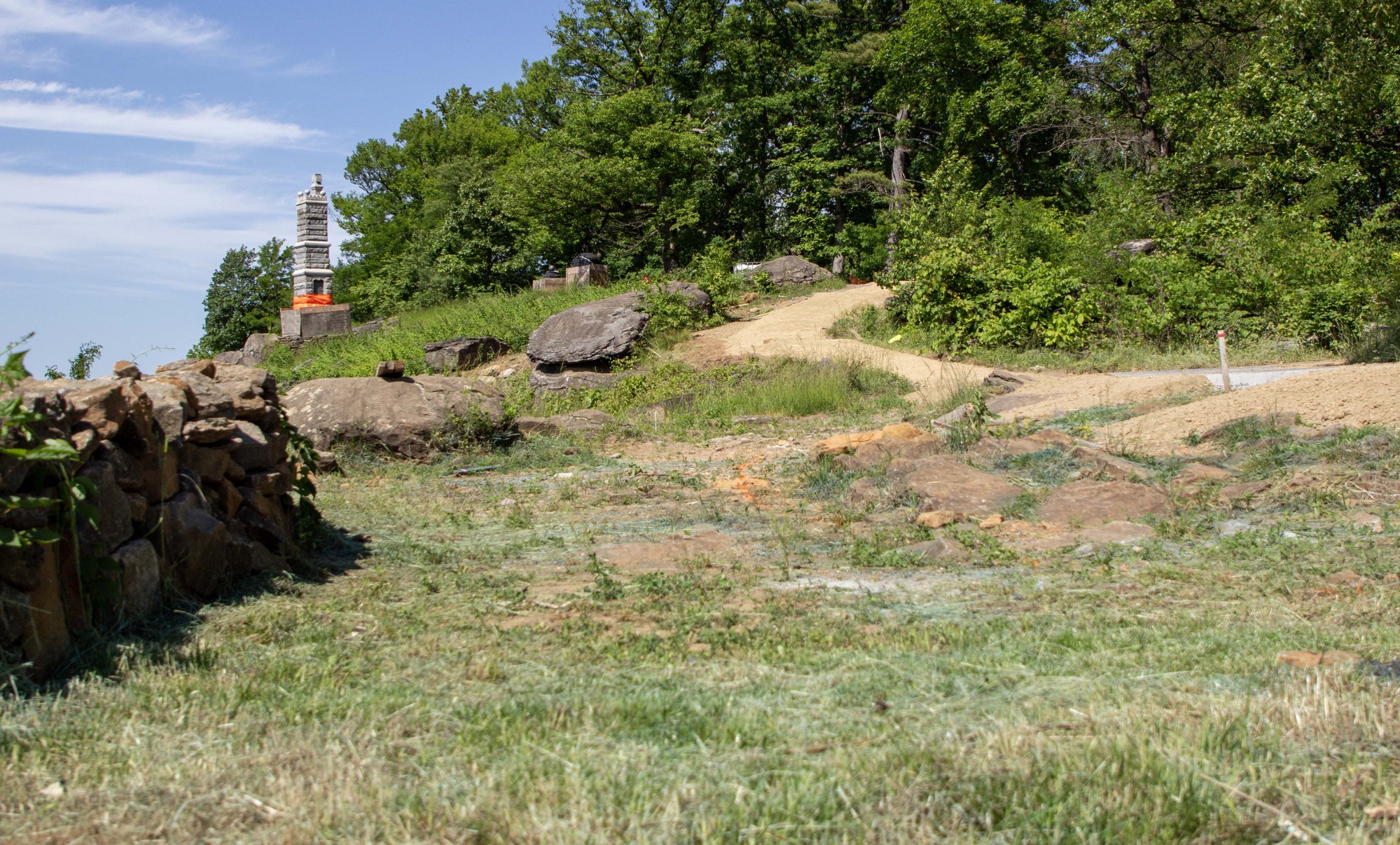 A grassy and rocky clearing sits next to a stone wall with a monument, trees, large boulders, and a brown path in the background.
