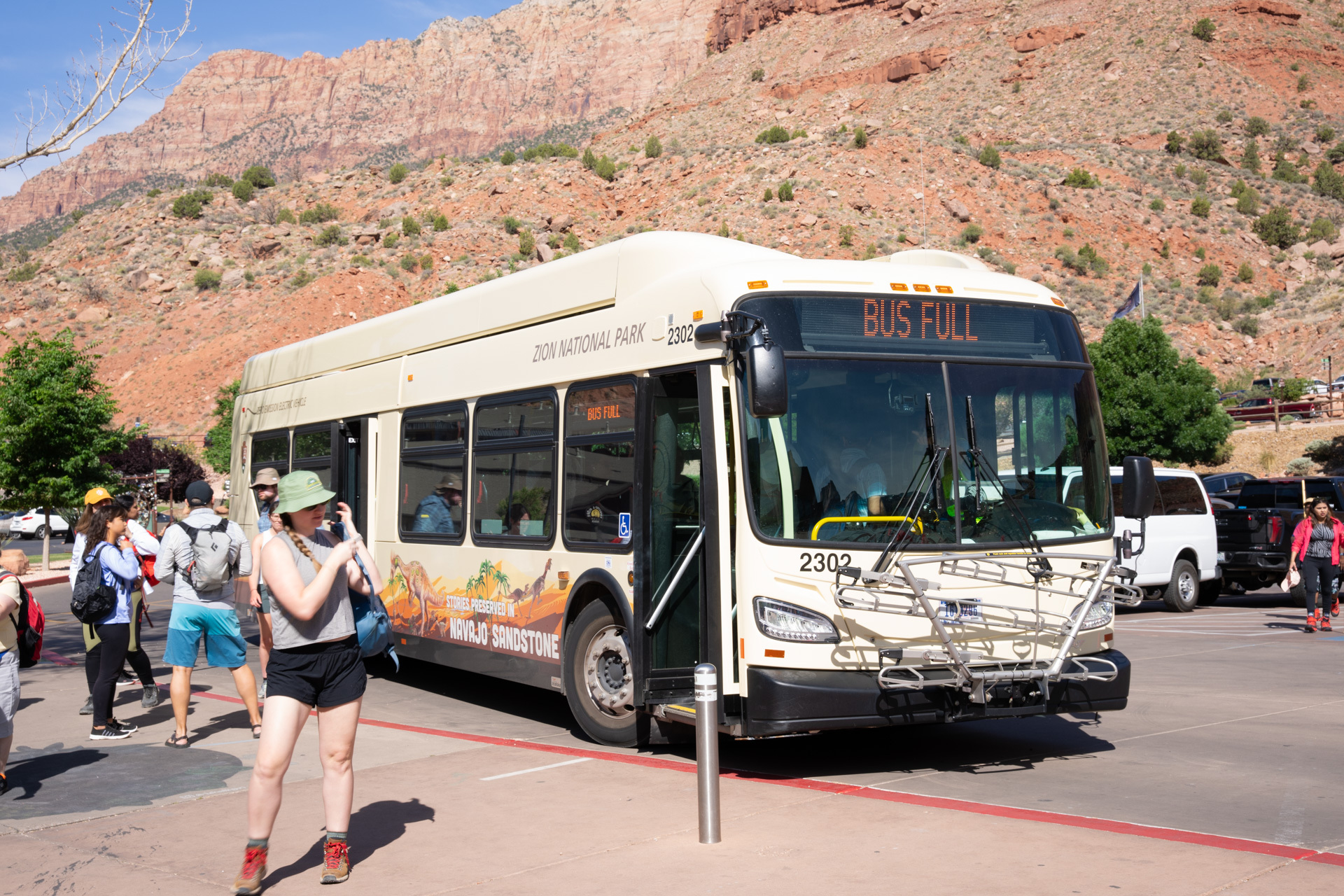 A white shuttle bus is parked alongside a curb. The digital panel on front of the bus reads "Bus Full." Visitors step out into a plaza, which offers views of the surrounding canyon.