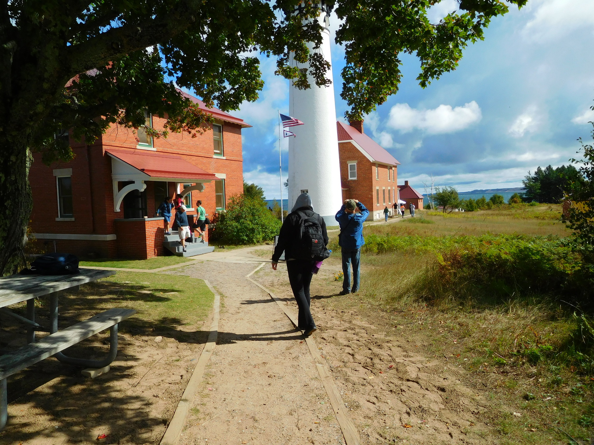 People on the grounds and porch of the Au Sable Light Station which consists of two brick two-story houses, a lighthouse, and a fog signal building.