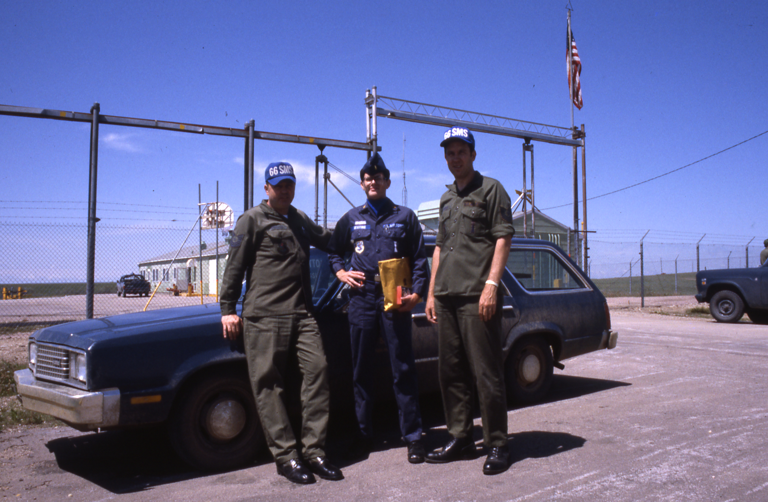 Three uniformed men stand in front of a fenced compound