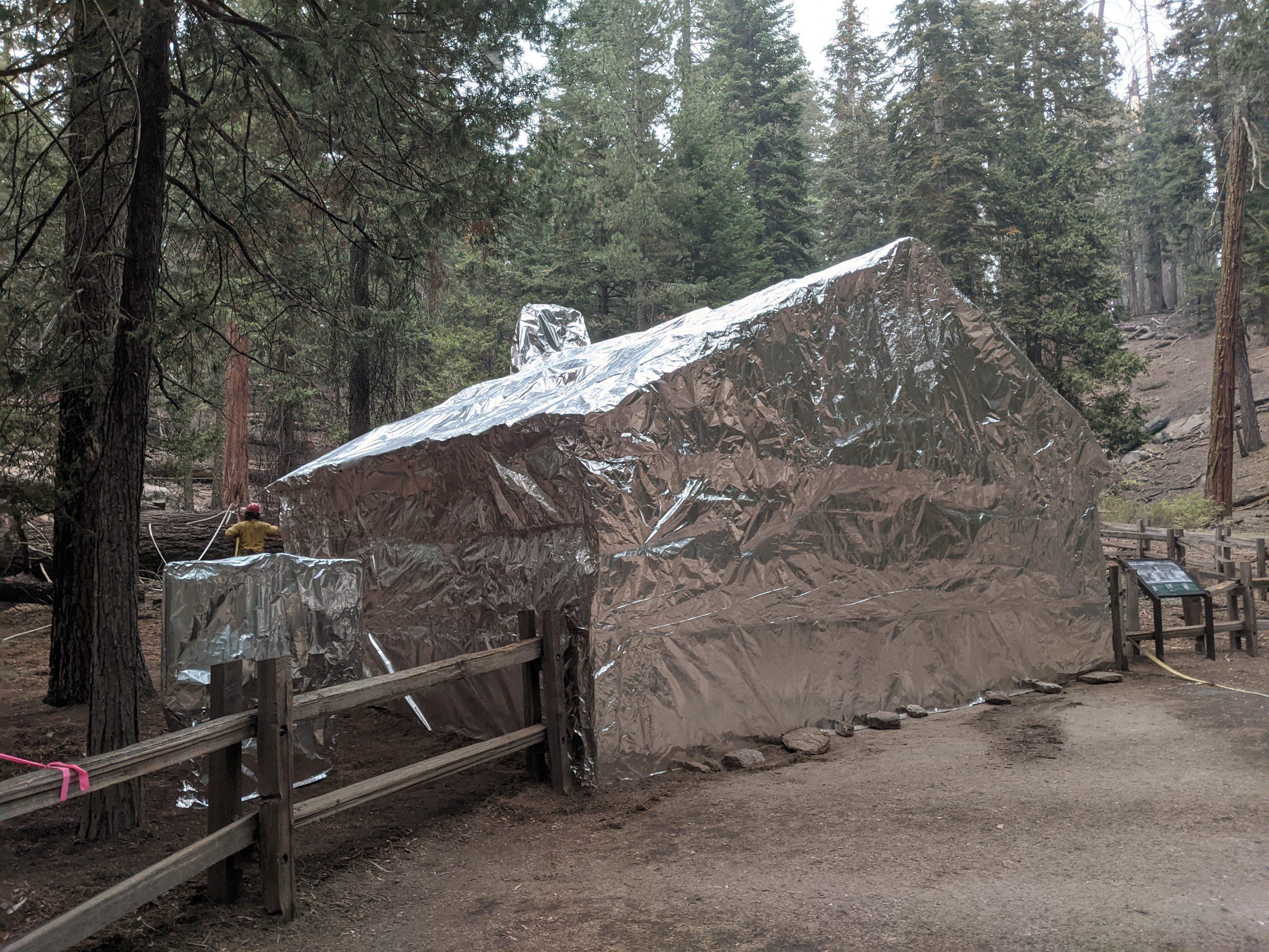 A small cabin and a sign, along a trail in a conifer forest, are entirely wrapped in shiny silver fire shelter material to protect them from an approaching wildfire. 