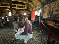 Two kids and an adult look into the blacksmith shop. In the back a blacksmith is working. 