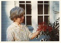 Color photograph of a woman with short grey hair and glasses from the waist up looking at a plant with red berries. There is a window and white brick wall behind her.