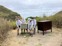 Four teens lift a large green metal box labeled with the number 