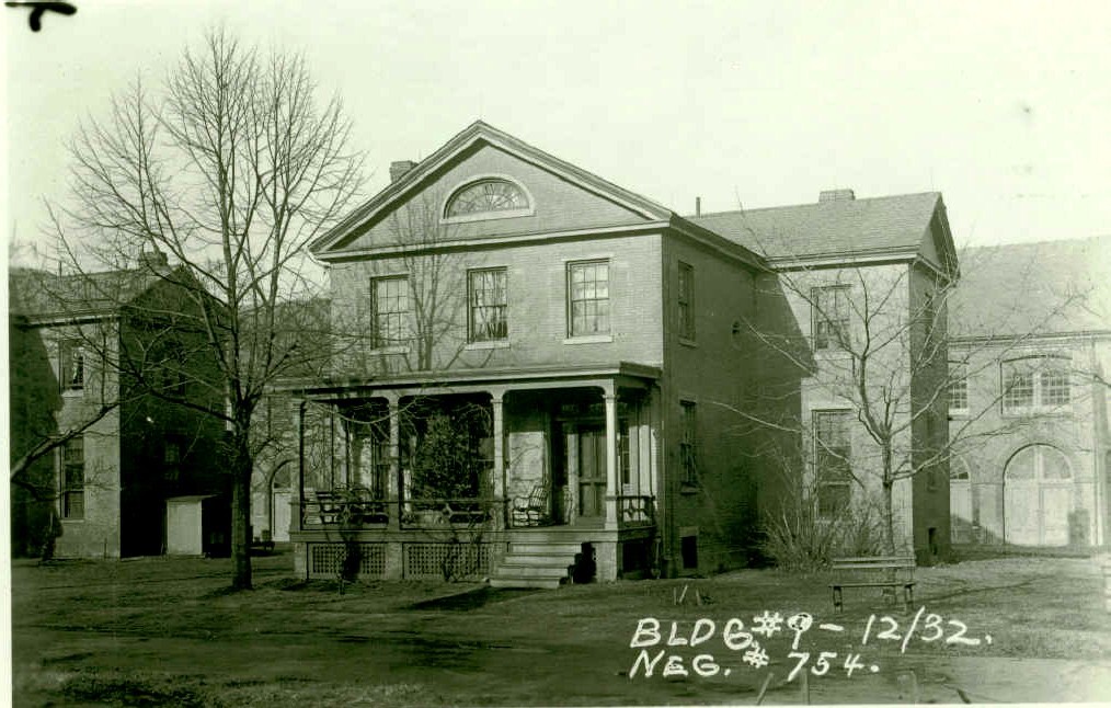 Black and white photo of a brick house with a front porch. Two trees out front. A small bench sits in front of the smaller tree on the right of the house. Writing on photo reads, “BLDG. 9- 12/32. NEG. 754.”