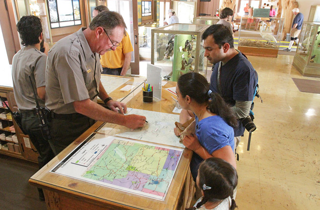 Male ranger is talking with a family and pointing to a  map on the counter top between them.