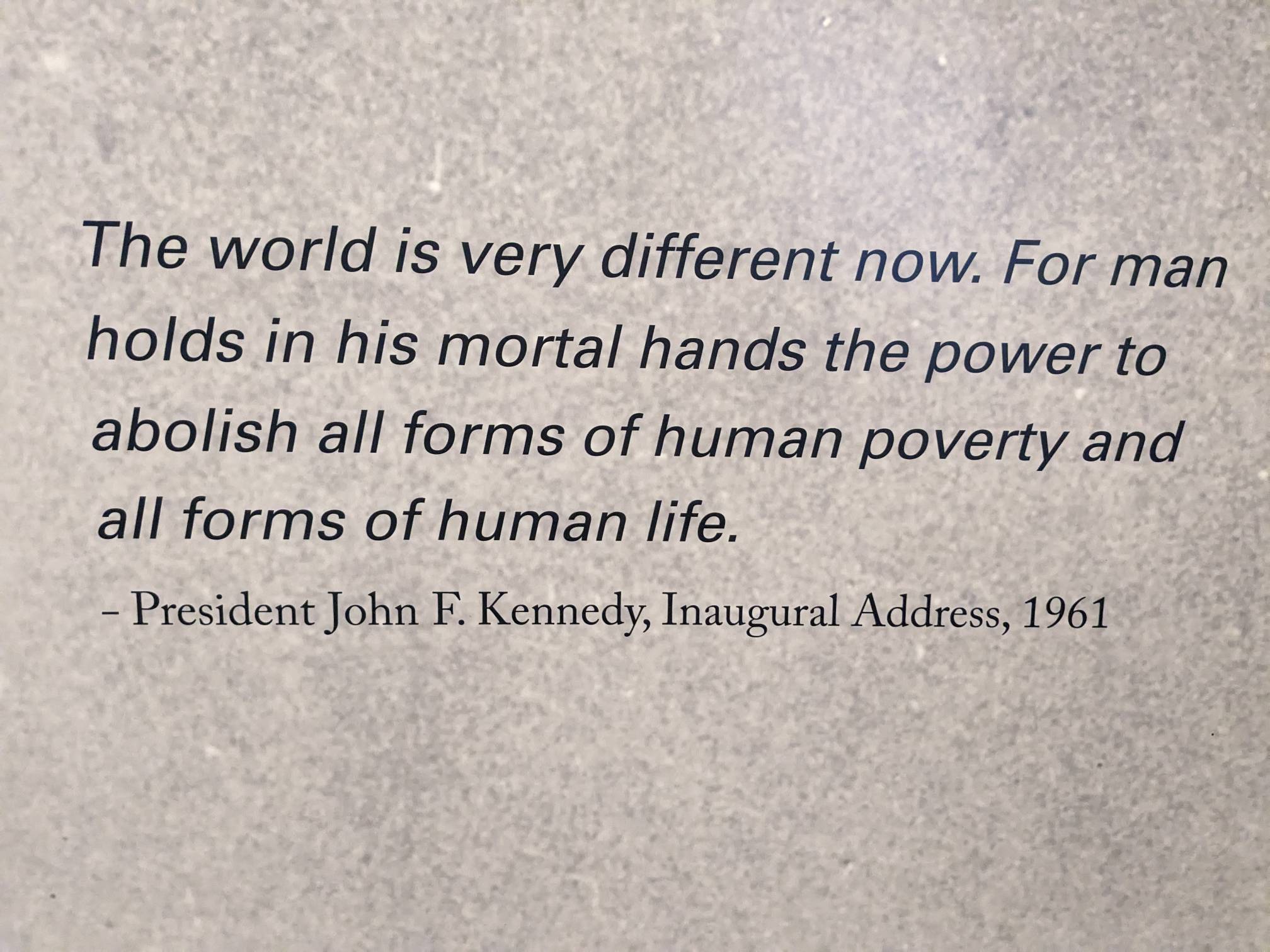 Black letters against a gray grainy background: "The world is very different now. For man holds in his mortal hands the power to abolish all forms of human poverty and all forms of human life. - President John F. Kennedy, Inaugural Address, 1961"