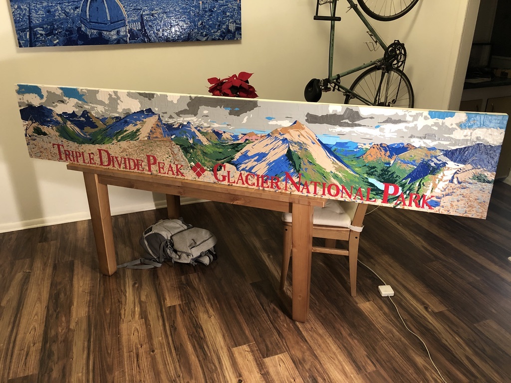 An image inside an artists studio of his duck tape and painters tape art piece of the view from Triple Divide Peak. This art piece features mountains stretching into the distance in the background, a lake in the background, rocky terrain in the foreground and clouds in the sky. The foreground also has the words "Triple Divide Peak. Glacier National Park" in red. The studio includes wood floors and a bicycle hanging on the wall.