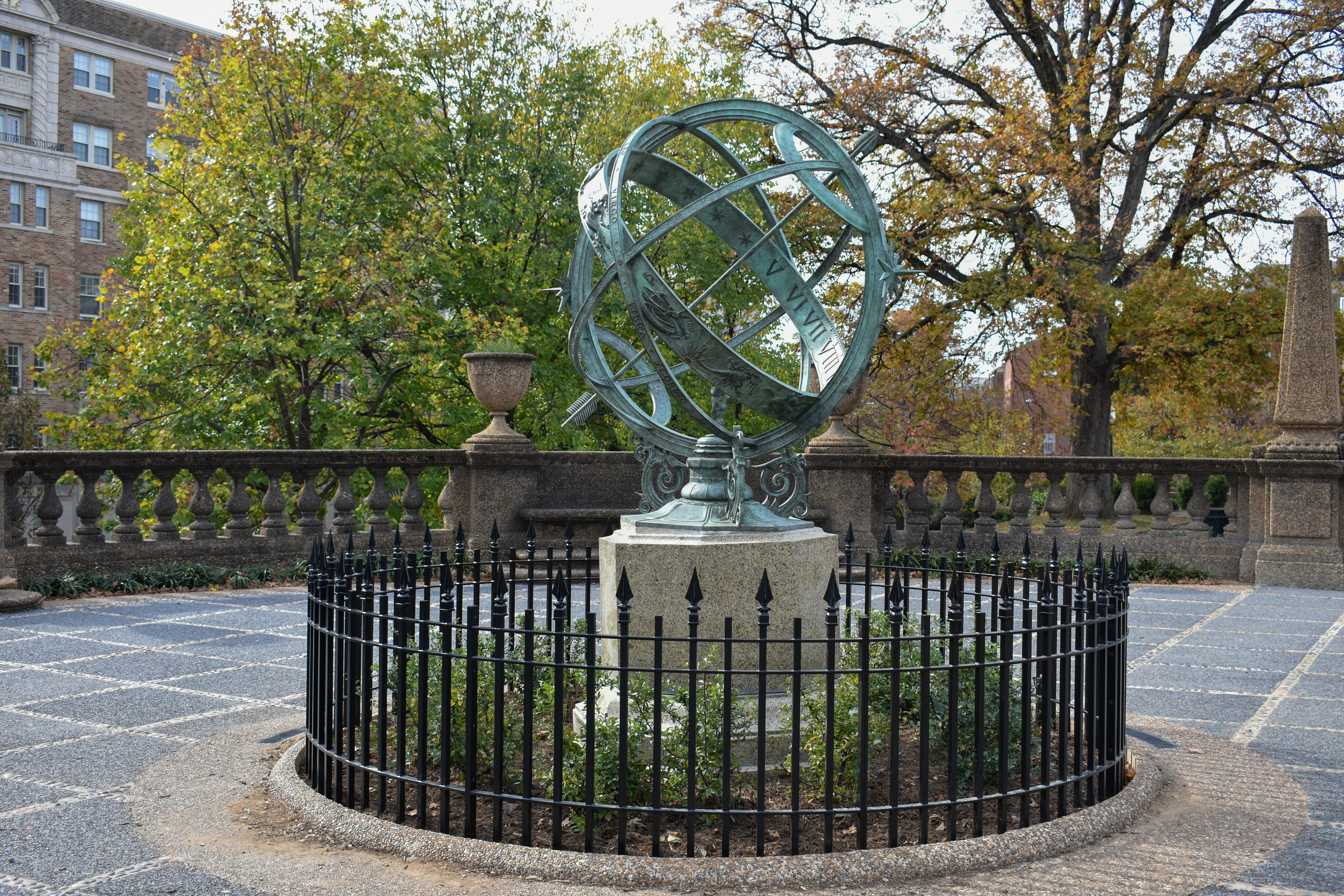 A green-tinted armillary sphere sculpture surrounded by bushes and pointed iron fence. It stands in front of fall trees and buildings in the distance.