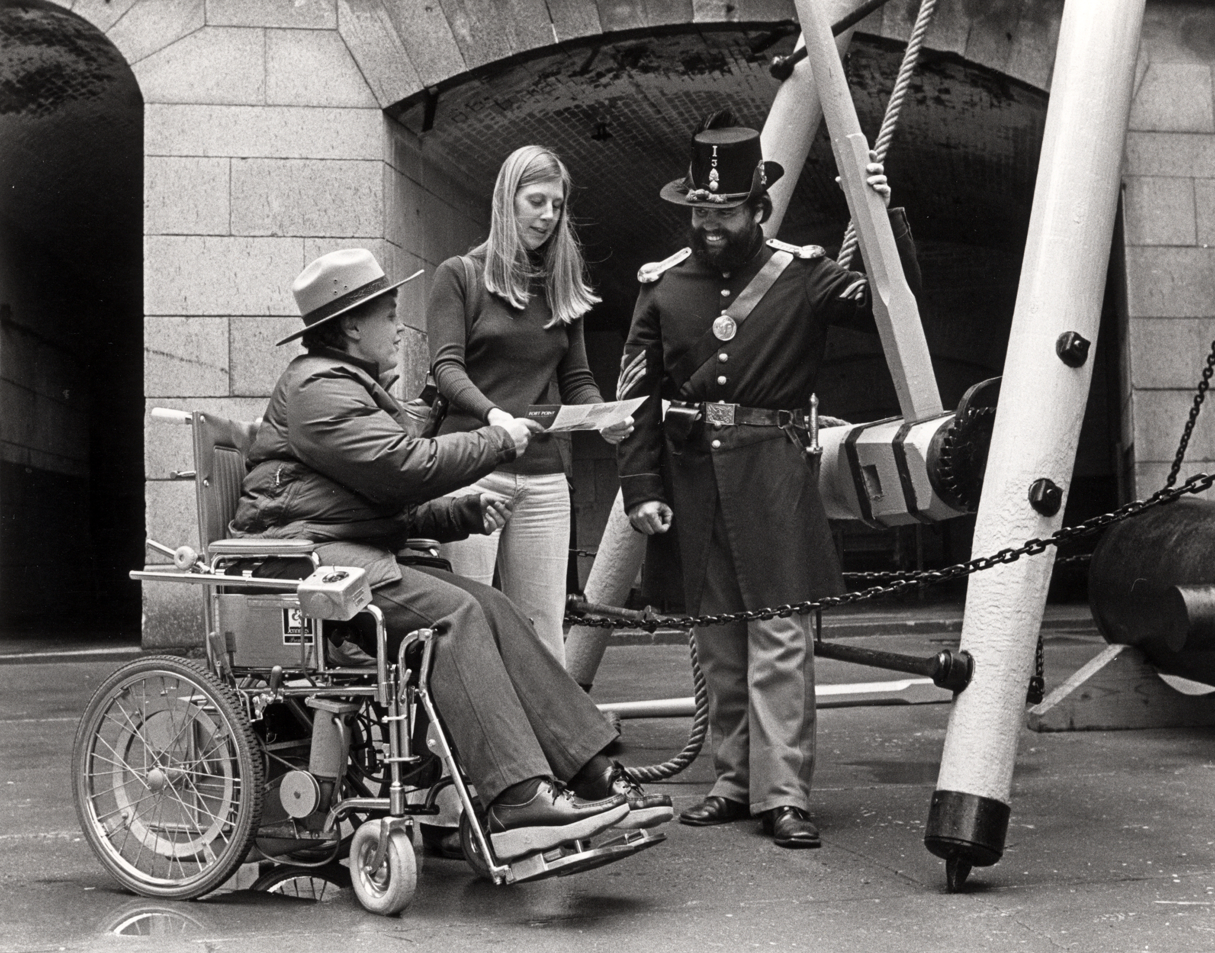 Vicky White in a wheelchair wears the NPS uniform and broad-brimmed hat as she discusses a brochure with a woman as a man in period military clothing looks on. 